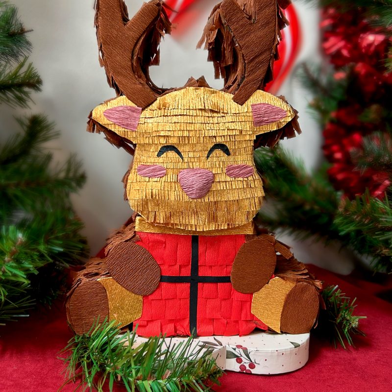 Piñata artisanale représentant un petit renne doré portant un cadeau rouge, dans un décor festif de Noël.